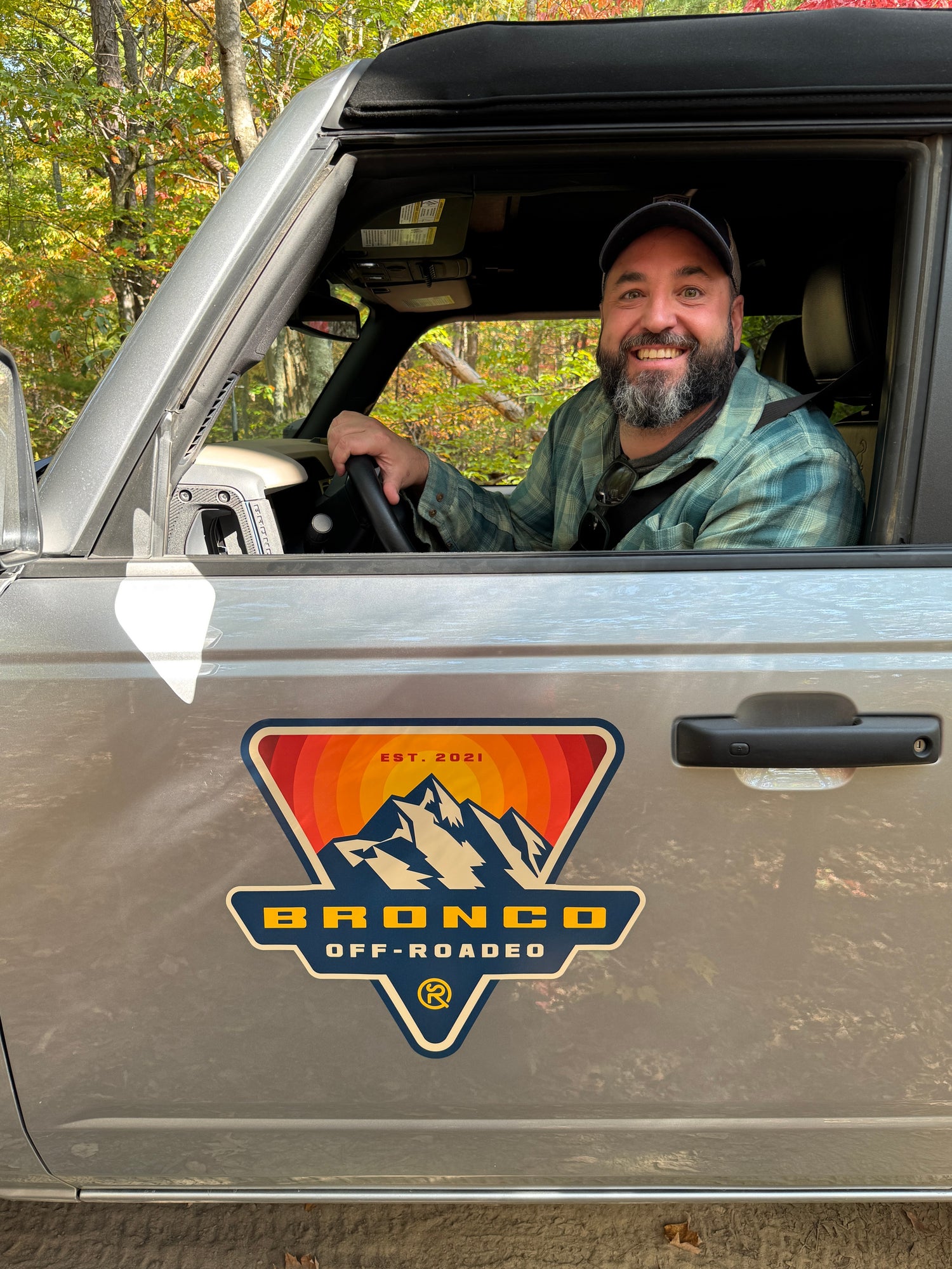 Man sitting in a vehicle with a Bronco off-road decal, surrounded by autumn foliage.